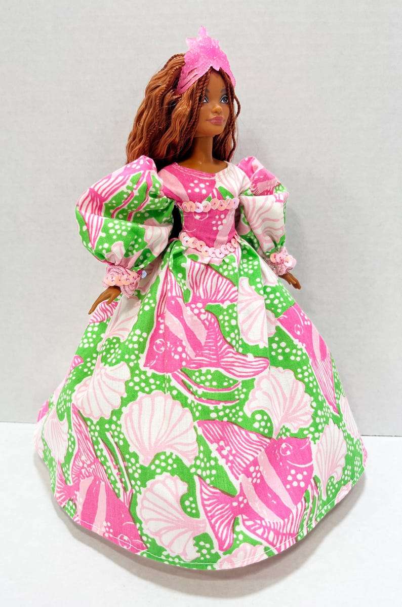 May include: A doll wearing a pink and green patterned dress with a matching pink tiara. The dress features a seashell and fish design. The doll has brown hair and is wearing a pink tiara. The dress has puffy sleeves and a full skirt.