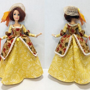 May include: A doll with brown hair wearing a straw hat with an orange ribbon and a dress featuring a yellow skirt with white pumpkin and leaf outlines, paired with a brown bodice patterned with red and orange maple leaves, holding a small brown basket.
