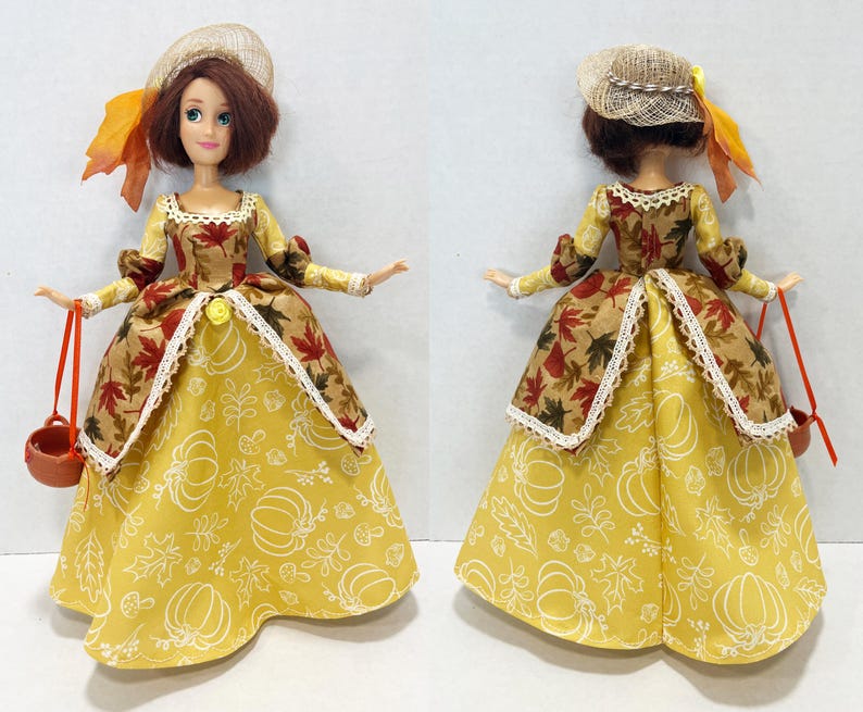 May include: A doll with brown hair wearing a straw hat with an orange ribbon and a dress featuring a yellow skirt with white pumpkin and leaf outlines, paired with a brown bodice patterned with red and orange maple leaves, holding a small brown basket.