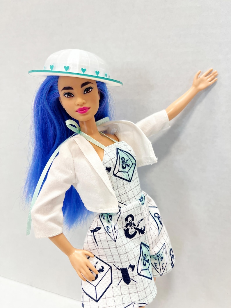 May include: A doll with long blue hair wearing a white hat with teal hearts, a white cropped jacket, and a white dress featuring a black grid pattern and various dark blue and teal gaming dice icons and symbols printed across the fabric.