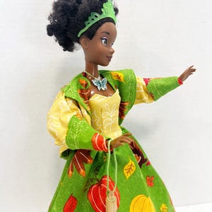 K&ouml;nnte beinhalten: Eine Puppe mit dunklem, lockigem Haar tr&auml;gt ein gr&uuml;nes Diadem, eine Schmetterlingskette und ein Kleid aus gr&uuml;nem Stoff mit einem Muster aus orangefarbenen und gelben K&uuml;rbissen und Herbstbl&auml;ttern, dazu eine gelbe Jacke und ein kleines Holzaccessoire.