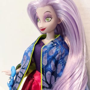 Pu&ograve; includere: Una bambola con lunghi capelli lisci color viola chiaro, occhi verdi e labbra rosse. La bambola indossa un top viola scuro, una giacca blu con un motivo a petali viola e bordi verde lime, e una gonna rossa. Un accessorio blu &egrave; visibile sul polso.