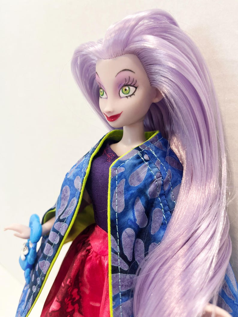 Pu&ograve; includere: Una bambola con lunghi capelli lisci color viola chiaro, occhi verdi e labbra rosse. La bambola indossa un top viola scuro, una giacca blu con un motivo a petali viola e bordi verde lime, e una gonna rossa. Un accessorio blu &egrave; visibile sul polso.
