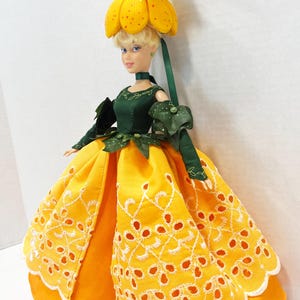 May include: A doll with blonde hair wearing a dark green bodice and a bright yellow skirt featuring white eyelet embroidery. The doll has a yellow flower-shaped hat with a small brown animal figure on top and green leaf accents at the waist.