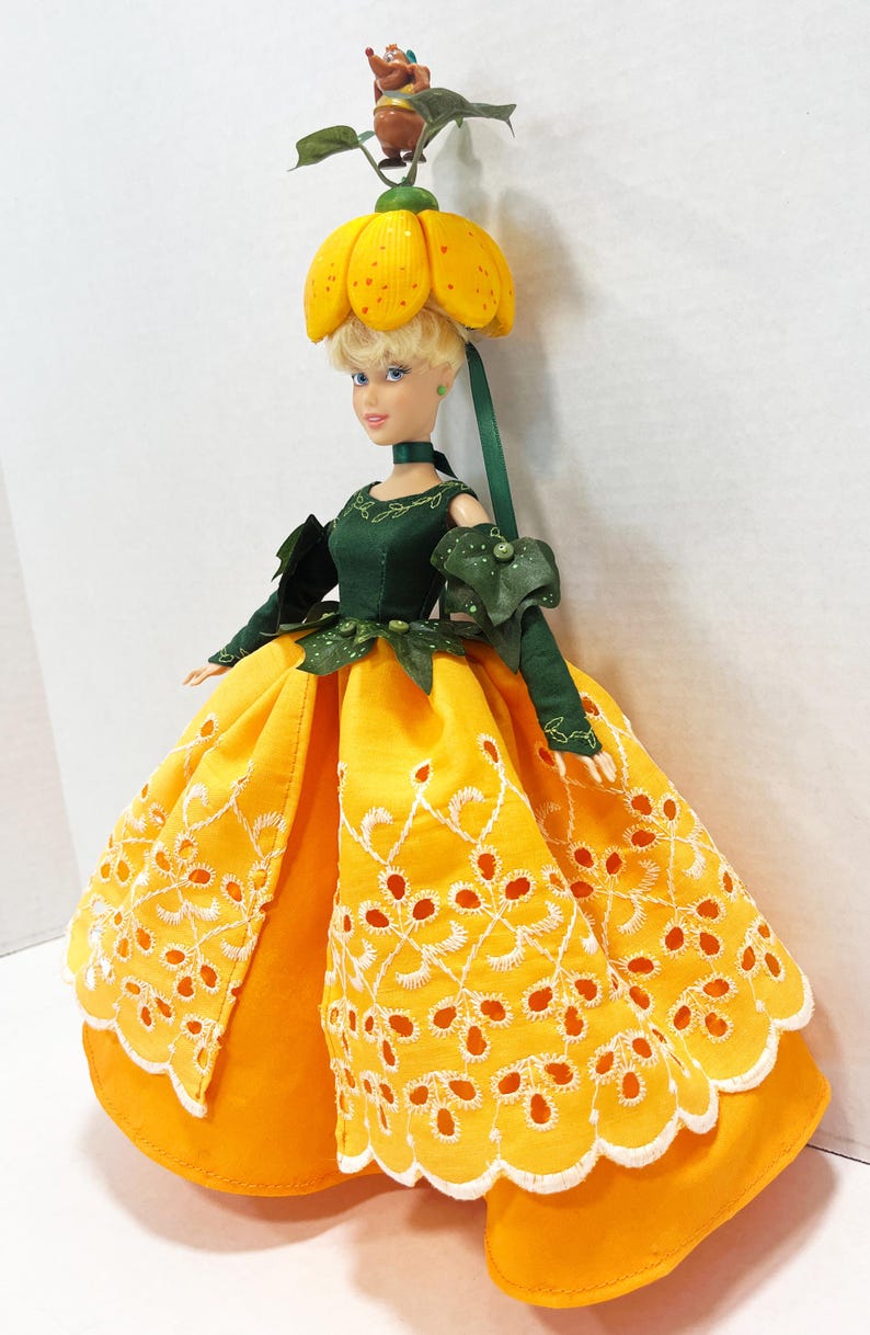 May include: A doll with blonde hair wearing a dark green bodice and a bright yellow skirt featuring white eyelet embroidery. The doll has a yellow flower-shaped hat with a small brown animal figure on top and green leaf accents at the waist.