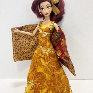 May include: A doll with dark red hair styled with a gold ribbon, wearing a yellow dress featuring a gold leaf pattern and gold leaf trim. The doll wears a reversible cape with a tan berry print on one side and a multi-colored autumn leaf print on the other.