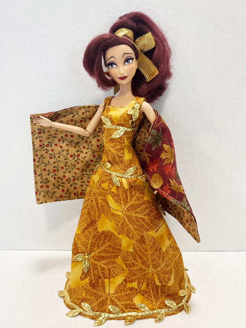 May include: A doll with dark red hair styled with a gold ribbon, wearing a yellow dress featuring a gold leaf pattern and gold leaf trim. The doll wears a reversible cape with a tan berry print on one side and a multi-colored autumn leaf print on the other.