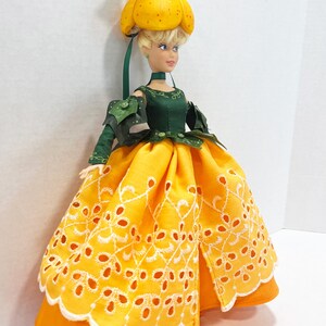 May include: A doll with blonde hair wearing a yellow pumpkin-shaped hat topped with a small brown creature and green leaves. The doll wears a dark green bodice with leaf-like sleeves and a bright yellow skirt featuring white eyelet embroidery.