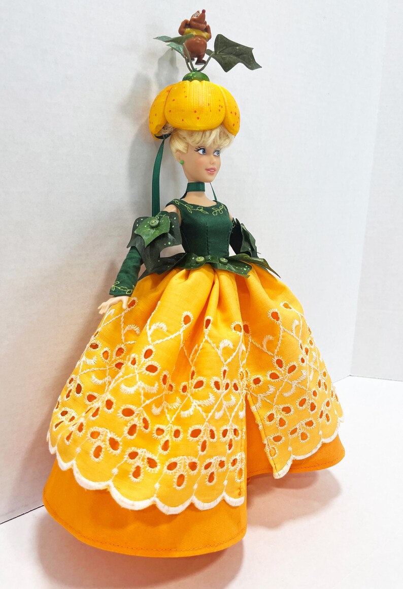 May include: A doll with blonde hair wearing a yellow pumpkin-shaped hat topped with a small brown creature and green leaves. The doll wears a dark green bodice with leaf-like sleeves and a bright yellow skirt featuring white eyelet embroidery.