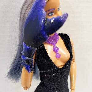 May include: A doll with blue and purple hair and makeup, wearing a black jumpsuit with glitter accents and a purple necklace. The doll has a large, dark blue and purple claw-like accessory over its arm.