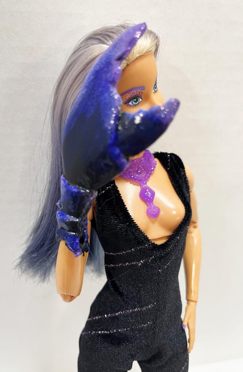 May include: A doll with blue and purple hair and makeup, wearing a black jumpsuit with glitter accents and a purple necklace. The doll has a large, dark blue and purple claw-like accessory over its arm.