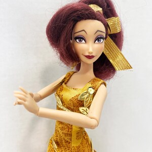 May include: A doll with dark reddish-purple hair styled in a high ponytail with a gold ribbon. The doll has purple eyes, dark eyebrows, and dark red lips. It wears a sleeveless gold dress featuring a leaf pattern and gold leaf-shaped accents.