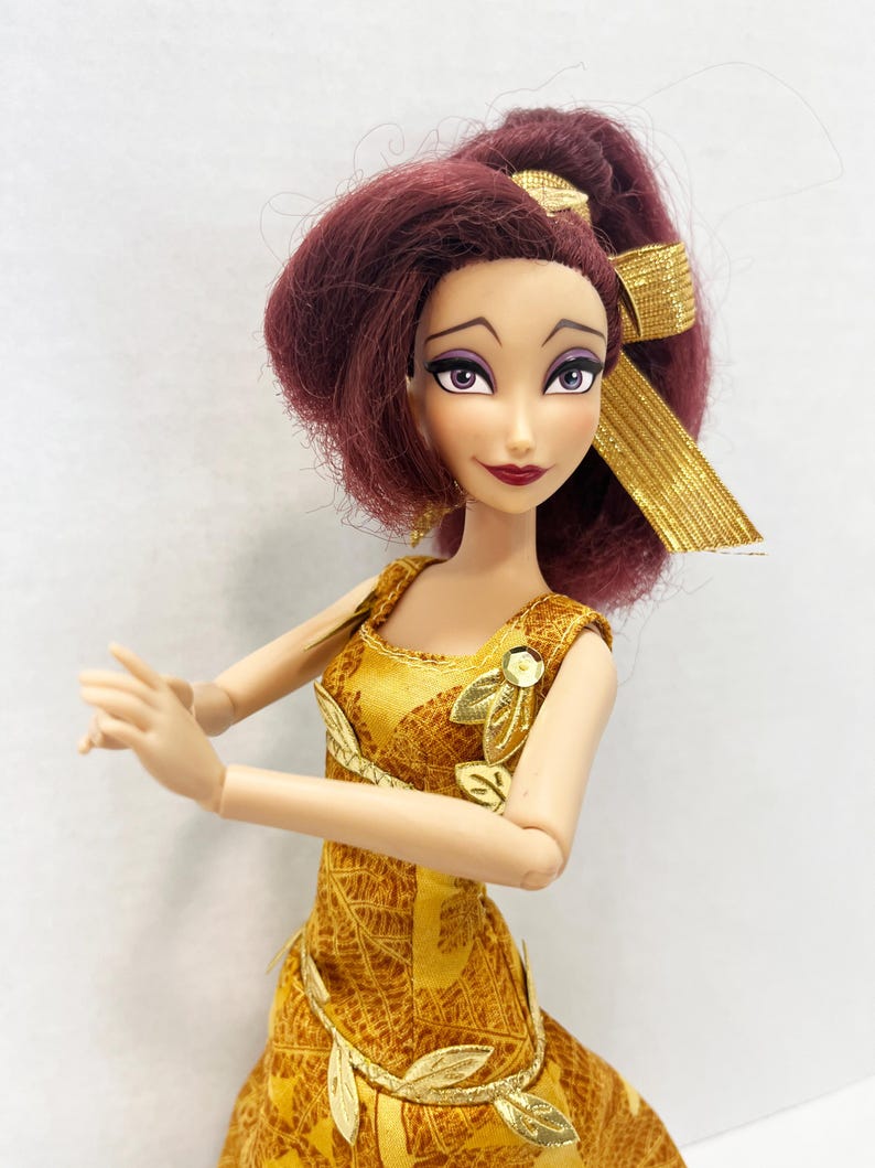 May include: A doll with dark reddish-purple hair styled in a high ponytail with a gold ribbon. The doll has purple eyes, dark eyebrows, and dark red lips. It wears a sleeveless gold dress featuring a leaf pattern and gold leaf-shaped accents.