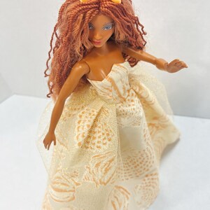 Puede incluir: Mu&ntilde;eca con piel morena, pelo largo casta&ntilde;o rojizo y una corona amarilla. La mu&ntilde;eca lleva un vestido crema sin tirantes con un estampado de pi&ntilde;a dorada. El vestido tiene una capa transparente. Los brazos de la mu&ntilde;eca est&aacute;n extendidos.