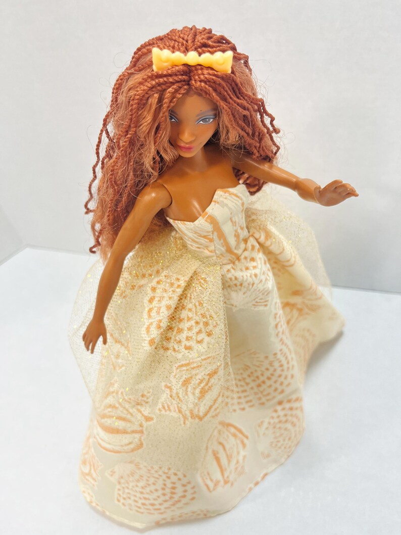 Puede incluir: Mu&ntilde;eca con piel morena, pelo largo casta&ntilde;o rojizo y una corona amarilla. La mu&ntilde;eca lleva un vestido crema sin tirantes con un estampado de pi&ntilde;a dorada. El vestido tiene una capa transparente. Los brazos de la mu&ntilde;eca est&aacute;n extendidos.