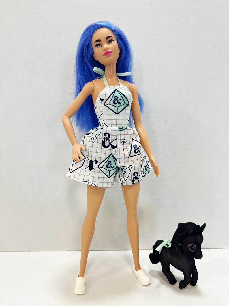 May include: A doll with long blue hair wearing a white halter-style romper featuring a black grid pattern and various gaming dice icons. The doll has light skin, pink lips, and white shoes. Beside the doll stands a small black toy horse figure.