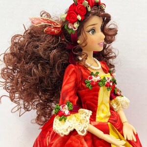 May include: A doll wearing a red rose-patterned dress with a yellow panel. The dress is adorned with floral accents and lace trim. The doll has brown curly hair, a red rose crown, and a pearl necklace.