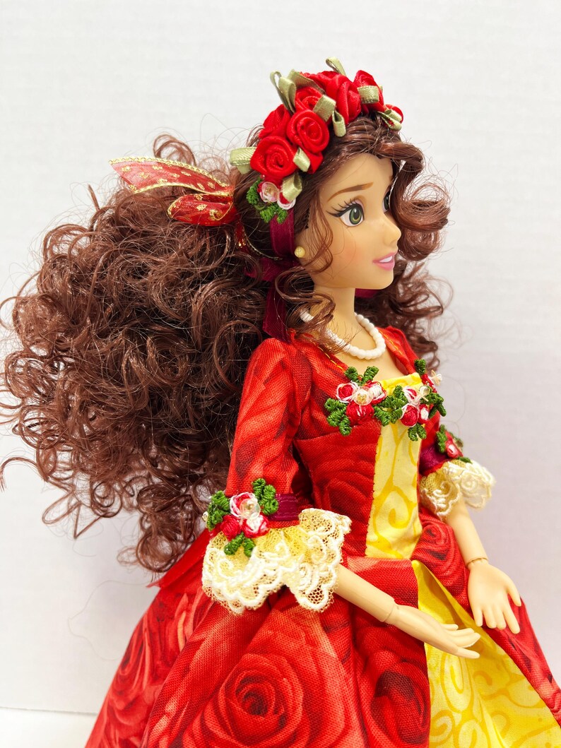 May include: A doll wearing a red rose-patterned dress with a yellow panel. The dress is adorned with floral accents and lace trim. The doll has brown curly hair, a red rose crown, and a pearl necklace.