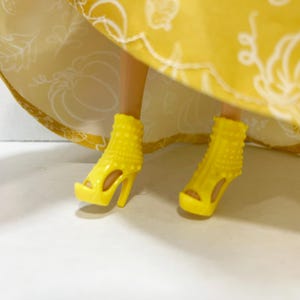 May include: A pair of bright yellow high-heeled platform sandals with a studded texture on the upper portion. The shoes are worn under a yellow skirt featuring a white line-art pattern of pumpkins and leaves on the fabric.