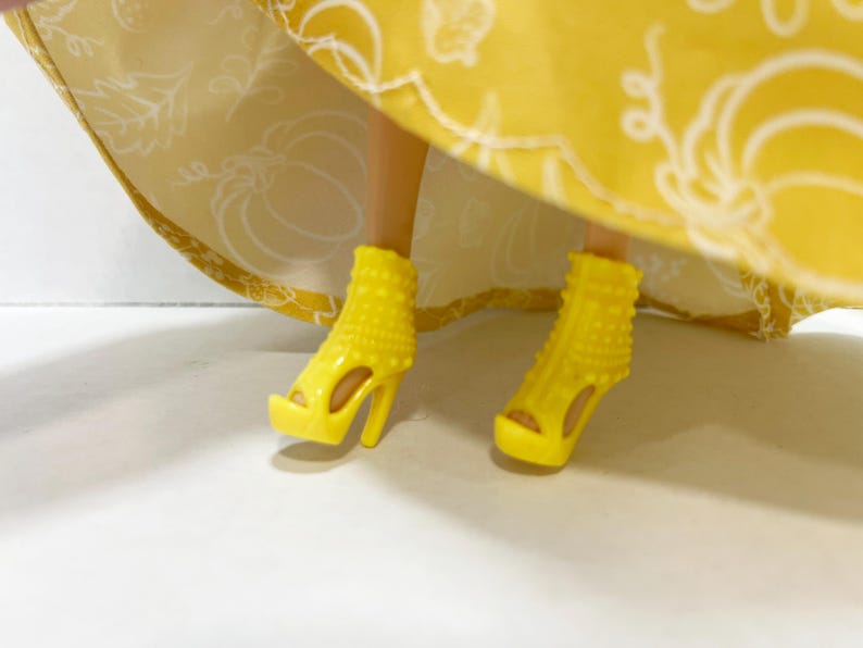 May include: A pair of bright yellow high-heeled platform sandals with a studded texture on the upper portion. The shoes are worn under a yellow skirt featuring a white line-art pattern of pumpkins and leaves on the fabric.