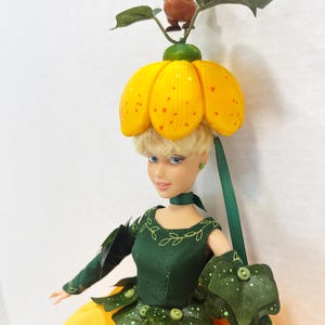 May include: A doll with blonde hair wearing a yellow pumpkin-shaped hat topped with a small brown mouse figure. The doll wears a dark green bodice with leaf-shaped accents, long green sleeves, and a bright yellow skirt with white embroidery.