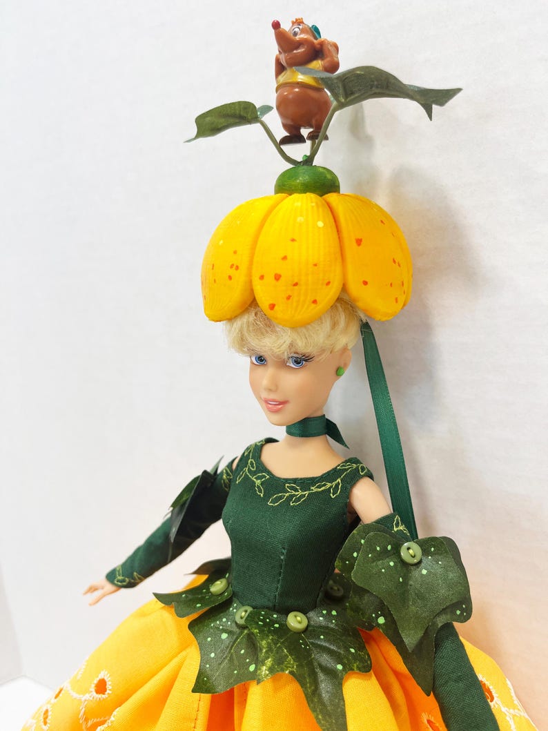 May include: A doll with blonde hair wearing a yellow pumpkin-shaped hat topped with a small brown mouse figure. The doll wears a dark green bodice with leaf-shaped accents, long green sleeves, and a bright yellow skirt with white embroidery.