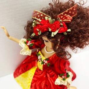 May include: A doll in a red rose-patterned dress with yellow accents. The doll has brown curly hair and wears a red rose-adorned headband with a matching bow. The dress features lace trim and a pearl necklace.