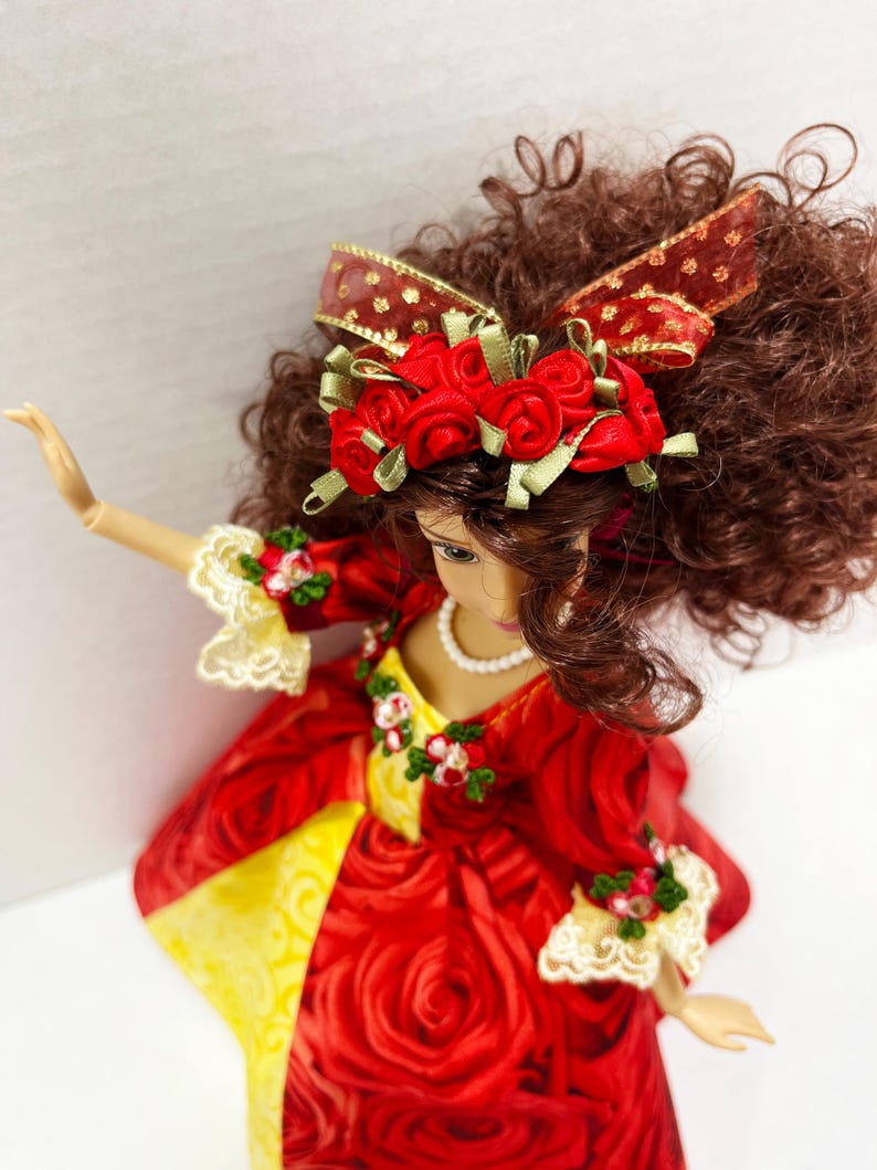 May include: A doll in a red rose-patterned dress with yellow accents. The doll has brown curly hair and wears a red rose-adorned headband with a matching bow. The dress features lace trim and a pearl necklace.
