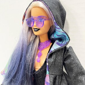 May include: A doll with long, lavender hair and purple-tinted sunglasses. The doll wears a black hooded jacket with silver glitter and a patterned lining. A purple necklace and black lipstick complete the look. The doll's hair is styled with a mix of silver and lavender.