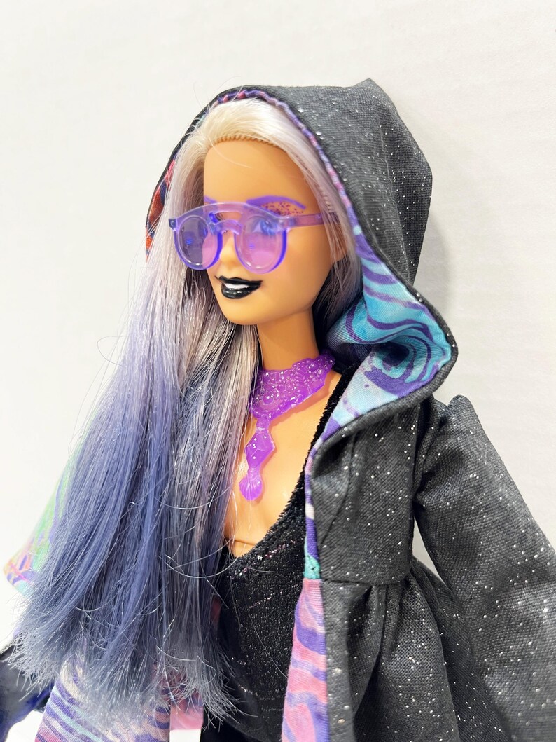 May include: A doll with long, lavender hair and purple-tinted sunglasses. The doll wears a black hooded jacket with silver glitter and a patterned lining. A purple necklace and black lipstick complete the look. The doll's hair is styled with a mix of silver and lavender.