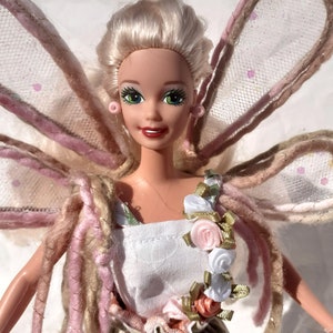 May include: A doll with blonde hair, green eyes, and pink earrings. The doll is wearing a white dress with a floral pattern and a garland of pink and white roses. It has pink and beige wings.