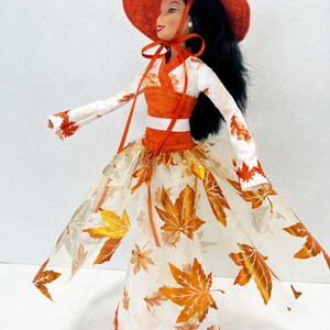 May include: A doll with long black hair wearing an orange wide-brimmed hat with a tie, a white long-sleeved top with orange leaf patterns, and a floor-length skirt featuring a sheer overlay decorated with metallic orange and gold maple leaf prints.