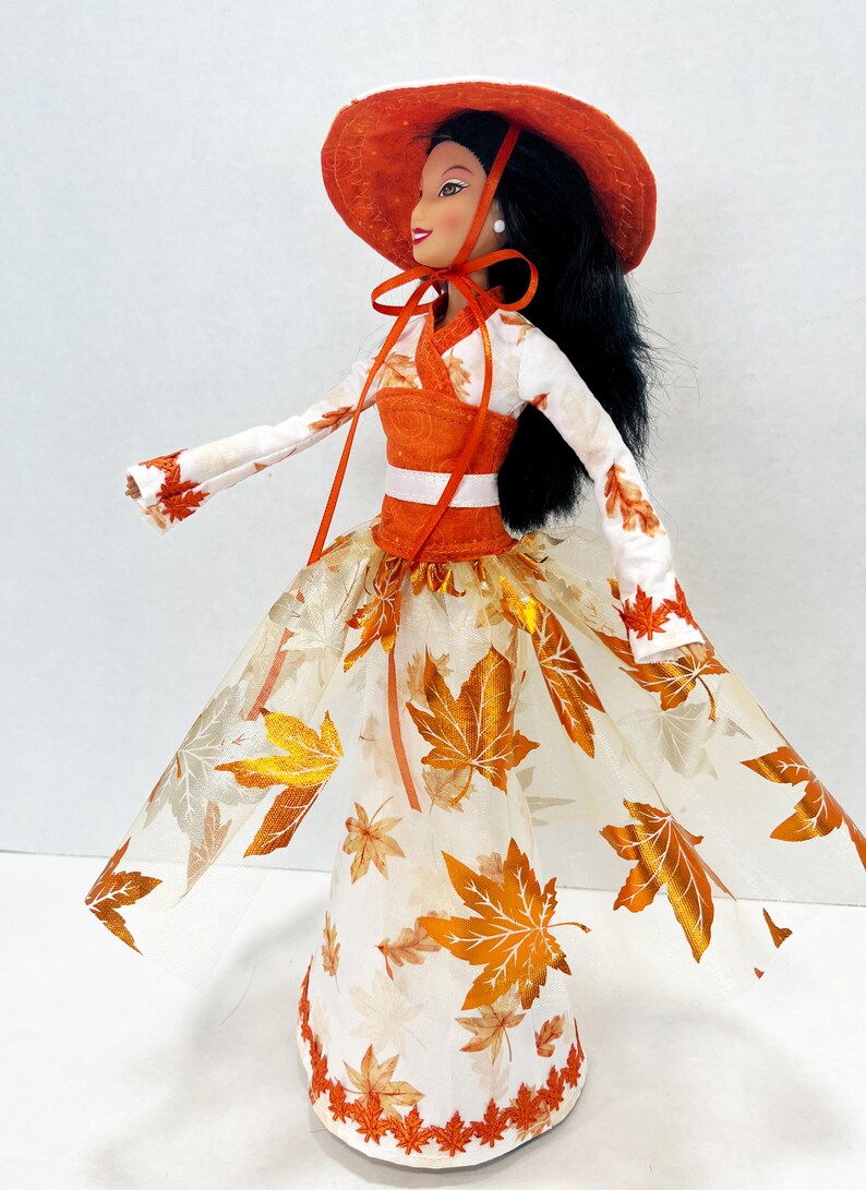 May include: A doll with long black hair wearing an orange wide-brimmed hat with a tie, a white long-sleeved top with orange leaf patterns, and a floor-length skirt featuring a sheer overlay decorated with metallic orange and gold maple leaf prints.