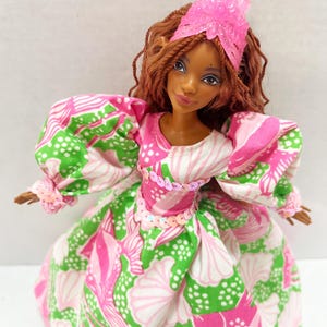 May include: A doll with brown hair and skin, wearing a pink and green patterned dress with puffy sleeves. The dress is adorned with sequins at the waist. A pink headpiece sits atop the doll's head.