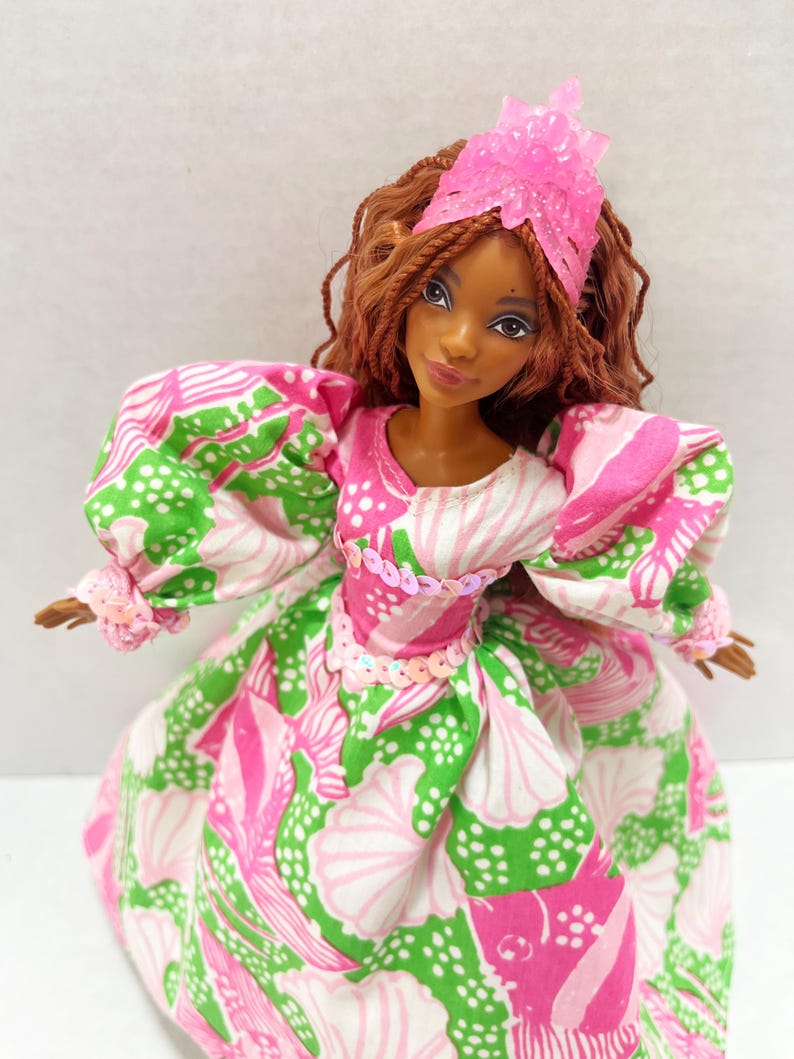 May include: A doll with brown hair and skin, wearing a pink and green patterned dress with puffy sleeves. The dress is adorned with sequins at the waist. A pink headpiece sits atop the doll's head.