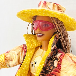 Puede incluir: Una mu&ntilde;eca con sombrero amarillo, gafas de sol rosas y chaqueta amarilla con estampado floral y de abejas. El atuendo de la mu&ntilde;eca incluye un top corto amarillo con un gr&aacute;fico de abeja. La mu&ntilde;eca tiene trenzas marrones y l&aacute;piz labial oscuro.