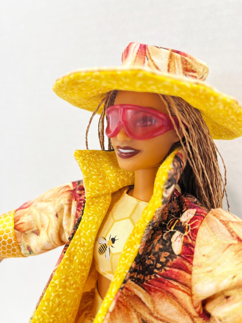 Puede incluir: Una mu&ntilde;eca con sombrero amarillo, gafas de sol rosas y chaqueta amarilla con estampado floral y de abejas. El atuendo de la mu&ntilde;eca incluye un top corto amarillo con un gr&aacute;fico de abeja. La mu&ntilde;eca tiene trenzas marrones y l&aacute;piz labial oscuro.