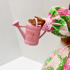May include: A small, pink watering can with glitter accents is held by a doll. The doll is wearing a colorful dress with a pink, green, and white floral pattern. The watering can has a handle and a spout.