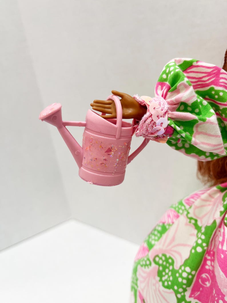 May include: A small, pink watering can with glitter accents is held by a doll. The doll is wearing a colorful dress with a pink, green, and white floral pattern. The watering can has a handle and a spout.