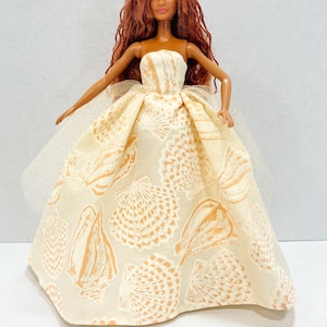 Puede incluir: Mu&ntilde;eca con un vestido crema sin tirantes con un estampado de conchas marinas. La mu&ntilde;eca tiene pelo largo color casta&ntilde;o rojizo y una peque&ntilde;a corona amarilla. Una peque&ntilde;a concha marina est&aacute; delante de la mu&ntilde;eca. El vestido tiene una capa de tul.