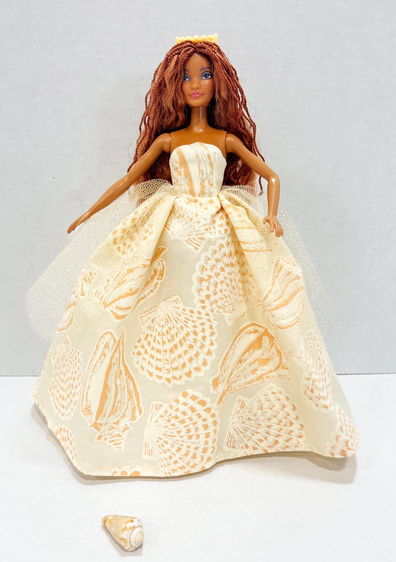 Puede incluir: Mu&ntilde;eca con un vestido crema sin tirantes con un estampado de conchas marinas. La mu&ntilde;eca tiene pelo largo color casta&ntilde;o rojizo y una peque&ntilde;a corona amarilla. Una peque&ntilde;a concha marina est&aacute; delante de la mu&ntilde;eca. El vestido tiene una capa de tul.