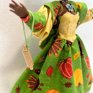 K&ouml;nnte beinhalten: Eine Puppe in einem gr&uuml;nen Kleid mit einem Muster aus K&uuml;rbissen, Truth&auml;hnen und Herbstbl&auml;ttern, kombiniert mit einem gelben Oberteil und einer passenden gr&uuml;nen Jacke. Ein kleines h&ouml;lzernes Nudelholz h&auml;ngt an einer gr&uuml;nen Schnur an der Hand, dazu ein Schmetterlingsanh&auml;nger.