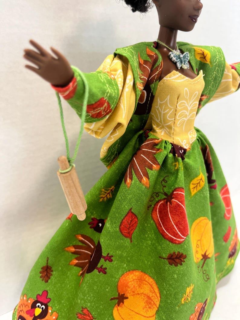 K&ouml;nnte beinhalten: Eine Puppe in einem gr&uuml;nen Kleid mit einem Muster aus K&uuml;rbissen, Truth&auml;hnen und Herbstbl&auml;ttern, kombiniert mit einem gelben Oberteil und einer passenden gr&uuml;nen Jacke. Ein kleines h&ouml;lzernes Nudelholz h&auml;ngt an einer gr&uuml;nen Schnur an der Hand, dazu ein Schmetterlingsanh&auml;nger.