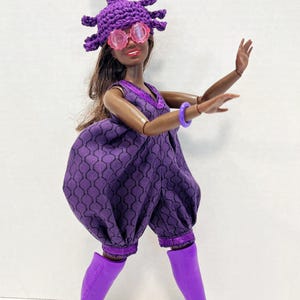 May include: A doll in a purple jumpsuit with a geometric pattern, knee-high boots, and a crocheted hat with a pink and green accent. The doll also wears pink-tinted glasses and a purple bracelet.