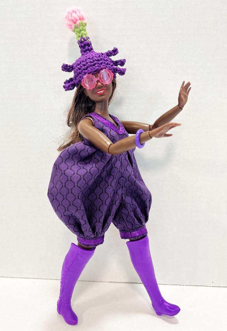 May include: A doll in a purple jumpsuit with a geometric pattern, knee-high boots, and a crocheted hat with a pink and green accent. The doll also wears pink-tinted glasses and a purple bracelet.