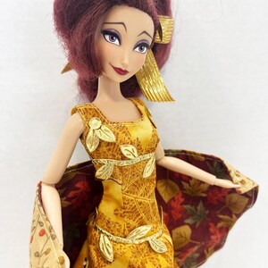 May include: A doll with deep red hair styled in a high ponytail with a gold ribbon. The doll wears a yellow sleeveless dress featuring a gold leaf pattern, accented with gold leaf appliqu&eacute;s at the neckline and waist, and a cape with a fall leaf print.