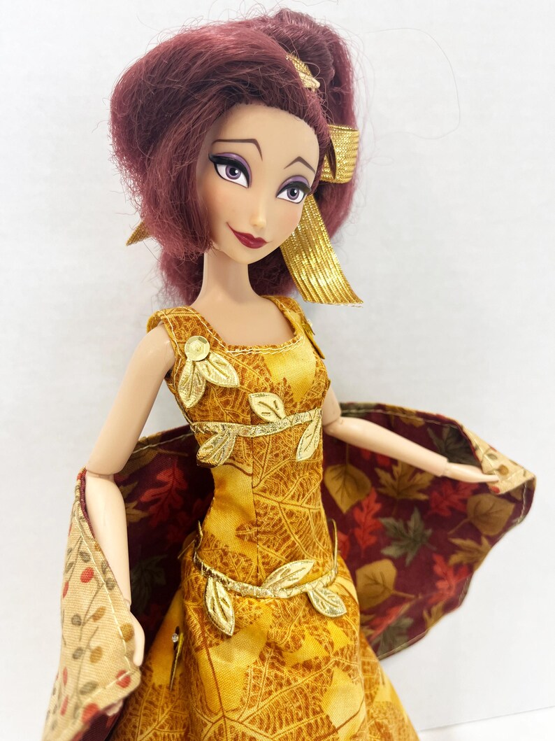 May include: A doll with deep red hair styled in a high ponytail with a gold ribbon. The doll wears a yellow sleeveless dress featuring a gold leaf pattern, accented with gold leaf appliqu&eacute;s at the neckline and waist, and a cape with a fall leaf print.