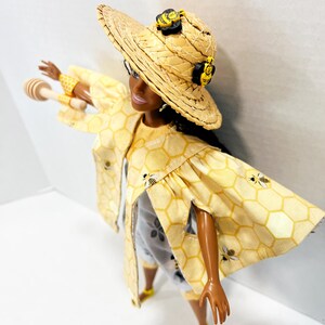 以下が含まれることがあります： 蜂のアクセントが付いた、黄色のハニカム模様の衣装を着た人形。 人形は、蜂の装飾が施された麦わら帽子、お揃いのジャケット、ショートパンツを着用しています。 人形は木製のハニーディッパーを持っており、黄色の靴を履いています。