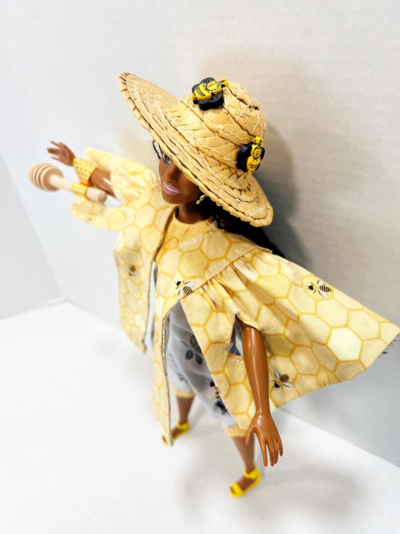 以下が含まれることがあります： 蜂のアクセントが付いた、黄色のハニカム模様の衣装を着た人形。 人形は、蜂の装飾が施された麦わら帽子、お揃いのジャケット、ショートパンツを着用しています。 人形は木製のハニーディッパーを持っており、黄色の靴を履いています。