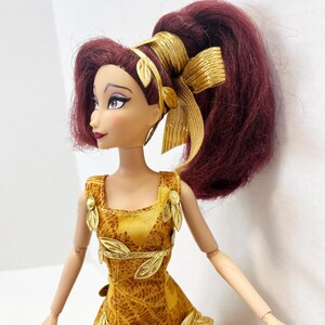 May include: A doll with dark reddish-purple hair styled in a high ponytail with a gold ribbon. The doll wears a yellow dress featuring a brown leaf pattern, accented with gold leaf-shaped embellishments on the bodice and waistline.